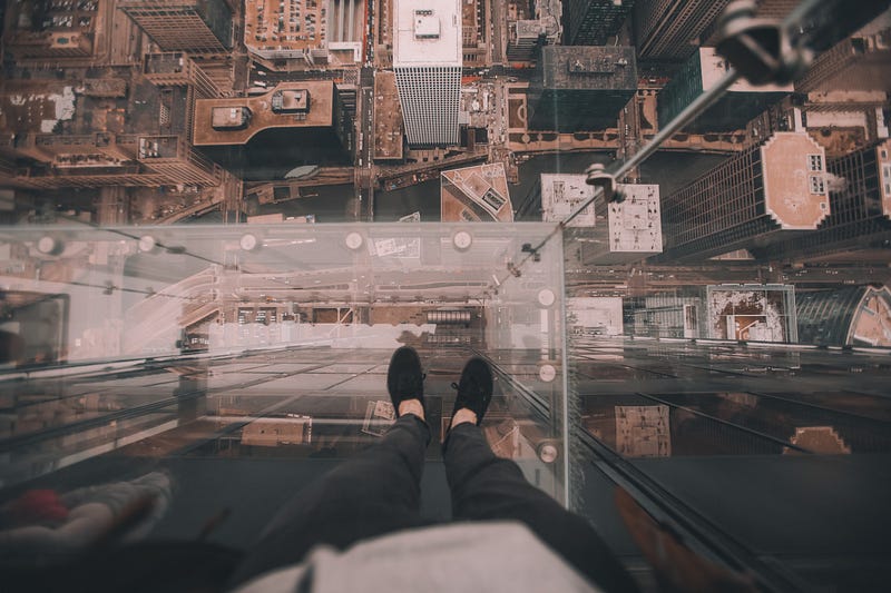 A person standing on a glass precipice on a tall building, looking down.