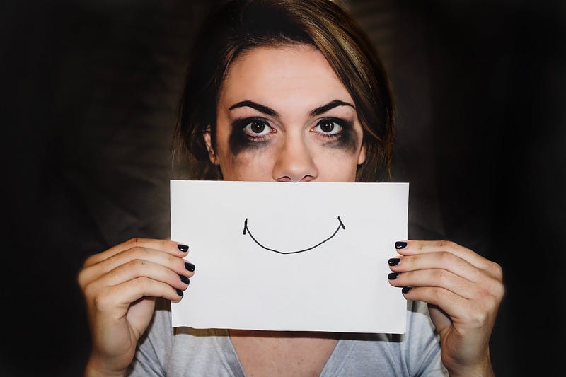 A person with running mascara and eyeshadow holding up a paper in front of their mouth with a drawn smile on it.